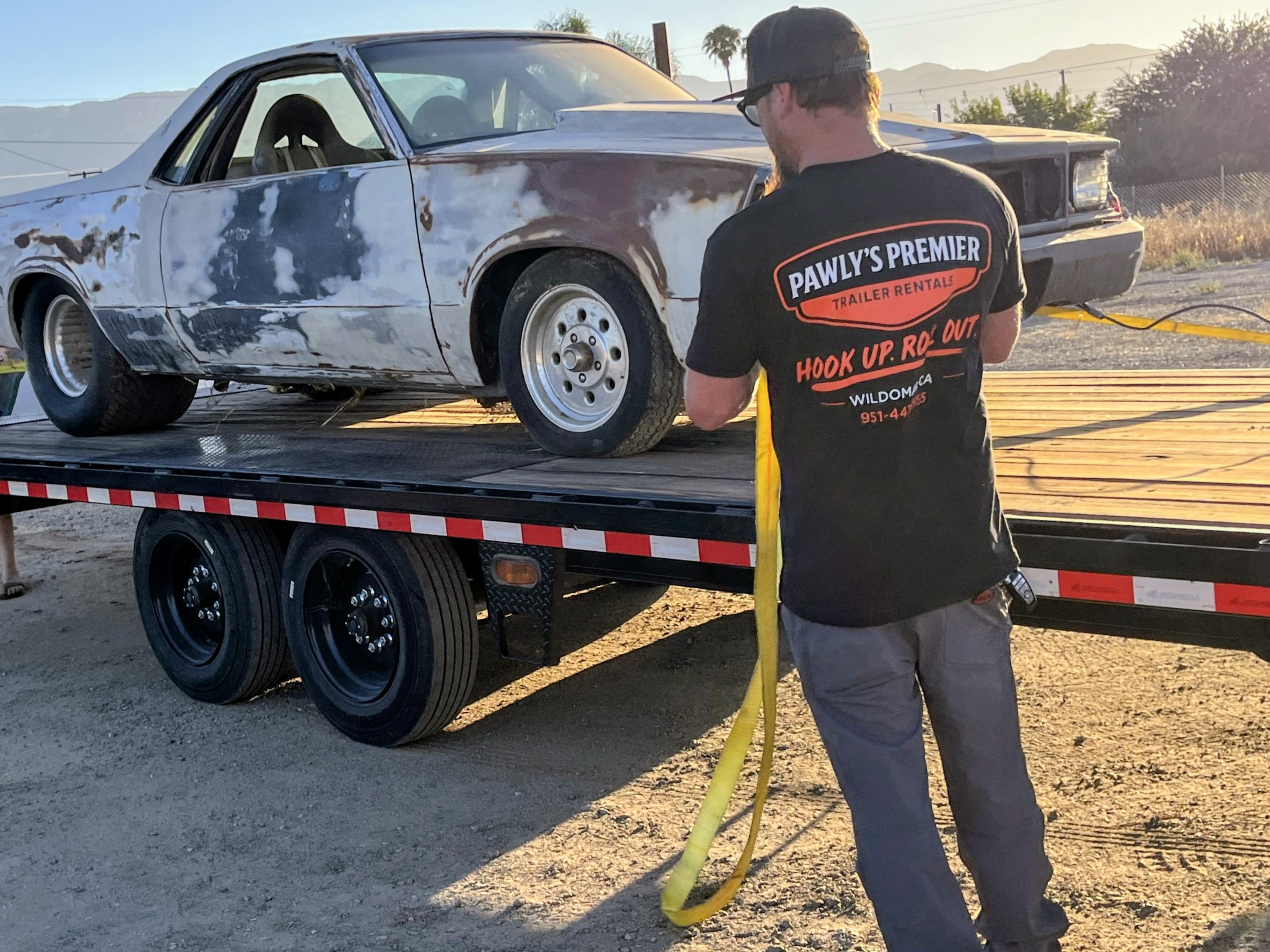 Man in Pawly's Premier shirt loading a project car onto a deckover tilt trailer at golden hour